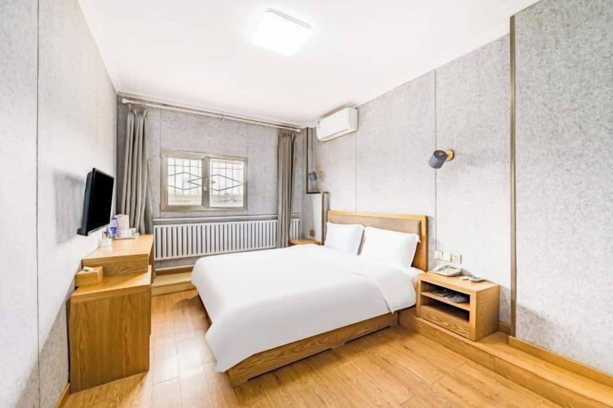 A hotel room with a bed, wooden bedside table, a desk with a chair, a window with curtains, and a radiator.