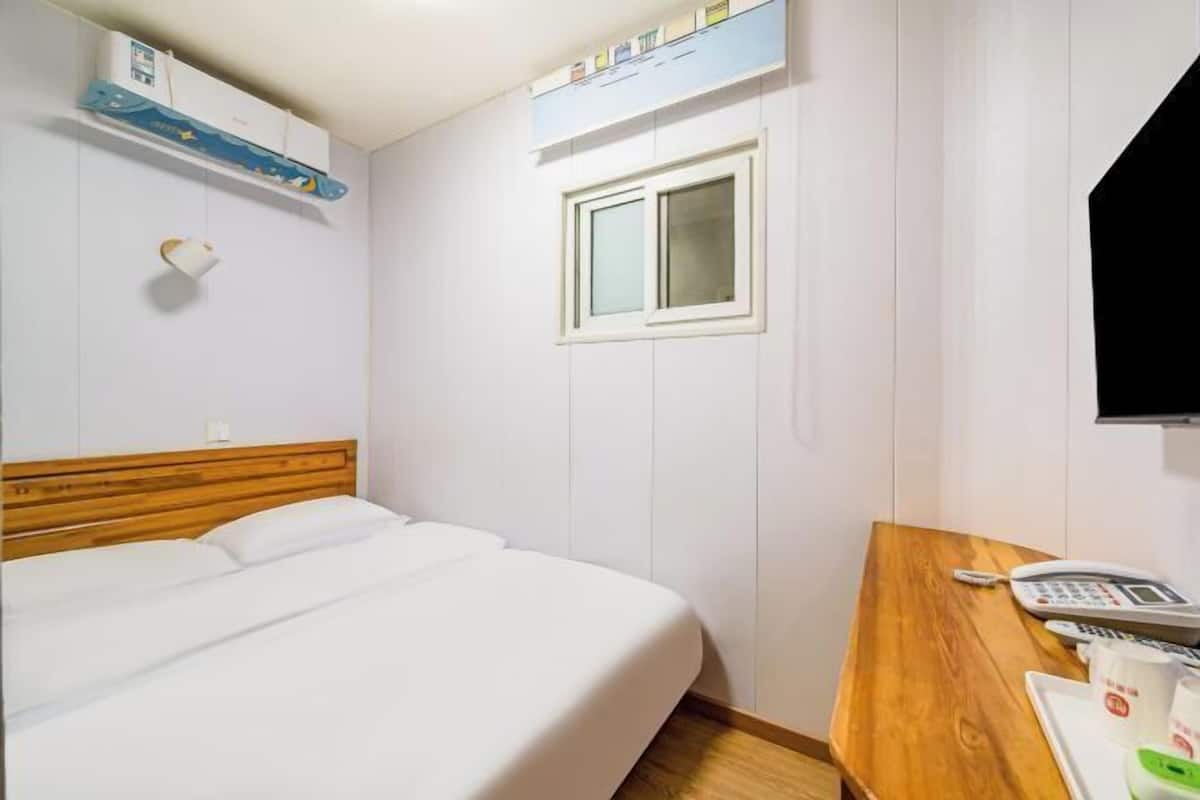 A hotel room with a bed, a wooden desk with a telephone, a window, and a wall-mounted air conditioner.