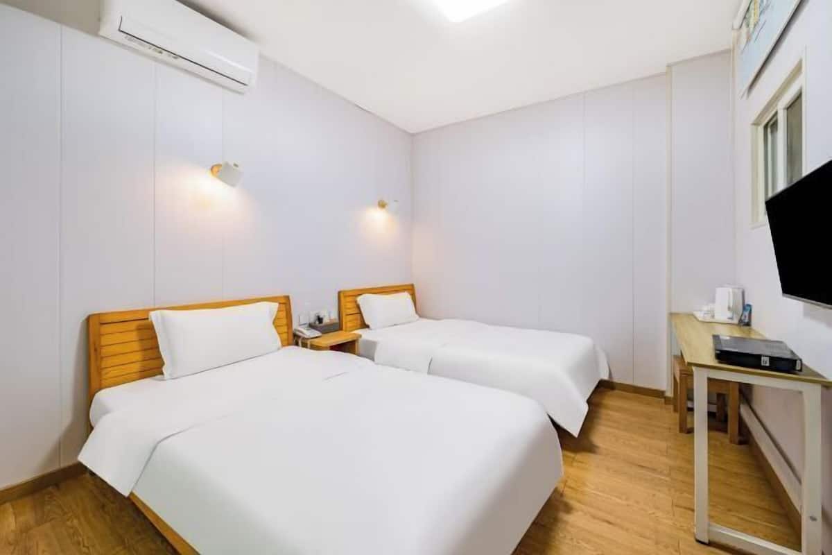 A hotel room with two beds, a wooden headboard, a small bedside table, a desk with a chair, a flat-screen TV, and an air conditioning unit.