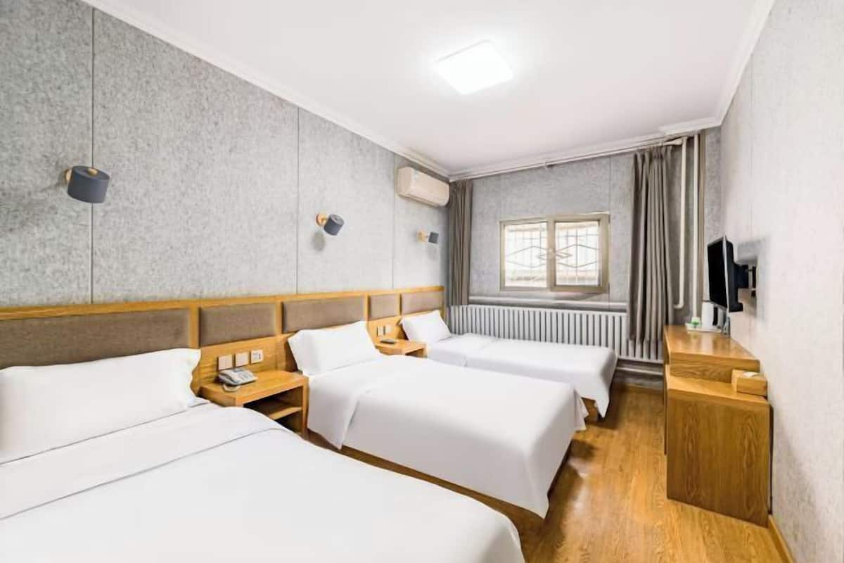 A hotel room with three beds, a wooden headboard, a radiator, a window with curtains, a TV, and a wooden desk.