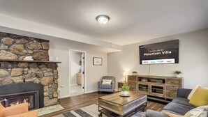 A living room with a stone fireplace, a flat-screen TV displaying a welcome message, a sofa, a coffee table, and a TV stand with a plant. - Mountain Villa! Scenic Getaway with Hot Tub (Cascade-Chipita Park)
