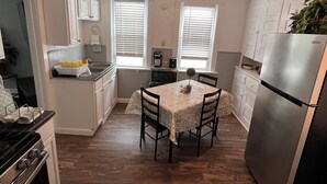 Una cucina compatta con mobili bianchi, un frigorifero in acciaio inossidabile, un tavolo con sedie e una ventola a soffitto.