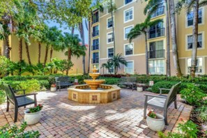 A courtyard with a central fountain, surrounded by benches and potted plants, with a multi-story building in the background. - Downtown WPB Condo at Cityplace (West Palm Beach)