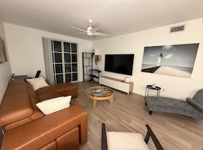 A modern living room with a brown leather sofa, a grey armchair, a round coffee table, and a flat-screen TV mounted on the wall. - Downtown WPB Condo at Cityplace (West Palm Beach)