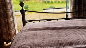 A bedroom with a large window, a bed with a patterned bedspread, and a view of a grassy field and distant buildings.