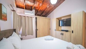 Une chambre avec un plafond en bois, un lit recouvert de draps blancs, un téléviseur fixé sur une commode en bois et une fenêtre avec des rideaux.
