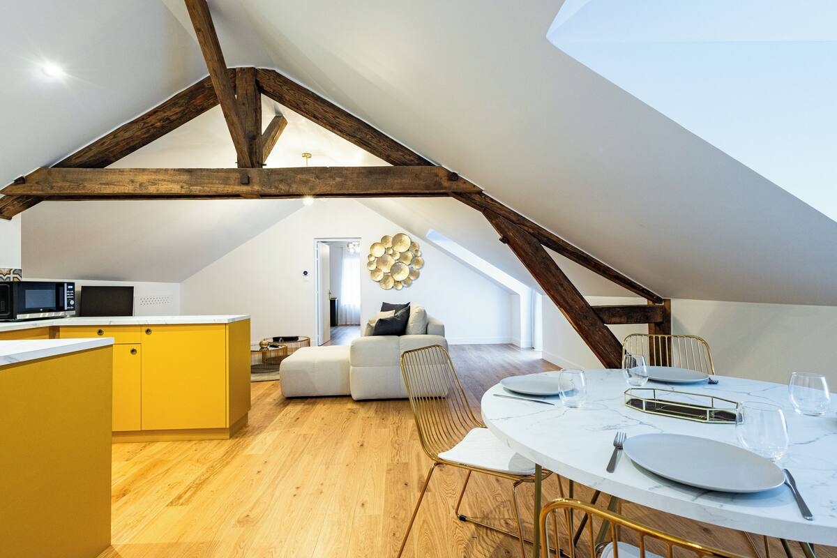 Ein modernes Loft mit freiliegenden Holzbalken, einer Küche mit gelben Schränken, einem Essbereich mit weißem Tisch und einer gemütlichen Sitzecke inklusive Sofa und dekorativem Wandbild.