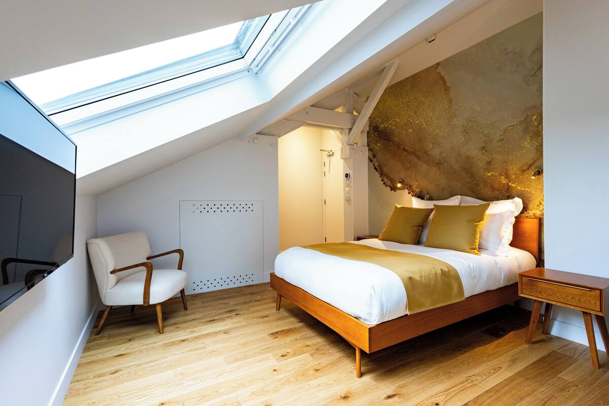 Ein modernes Schlafzimmer mit Dachfenster, einem Bett mit Holzrahmen, einem Sessel und einem Nachttisch.