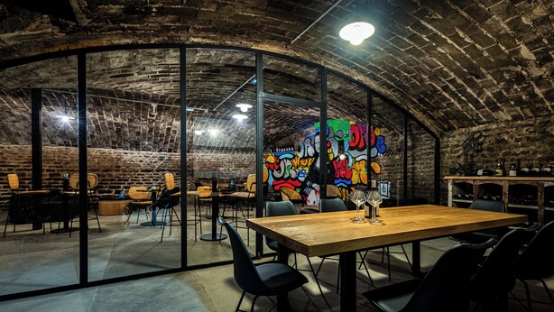 A rustic underground dining area with exposed brick walls, a graffiti mural, and a wooden table with chairs. - Solstice Beaune - Hotel et Terroir (Beaune)