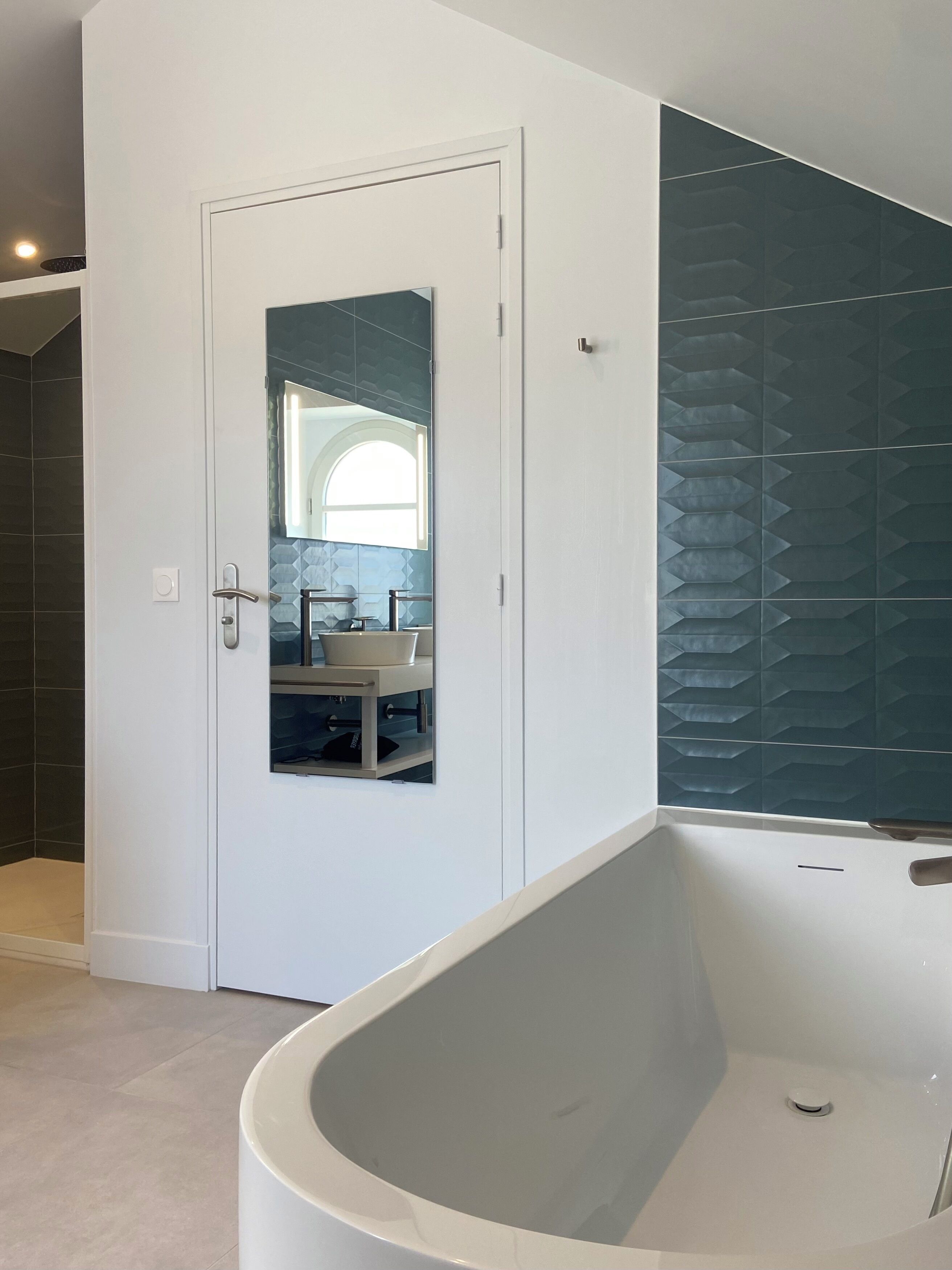 Ein Badezimmer mit einer weißen Badewanne, einer Tür inklusive Spiegel und einer gefliesten Wand.