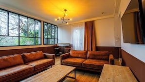 A modern living room with leather sofas, a wooden coffee table, and a dining area. Large windows offer a view of trees outside. - Solstice Beaune - Hotel et Terroir (Beaune)