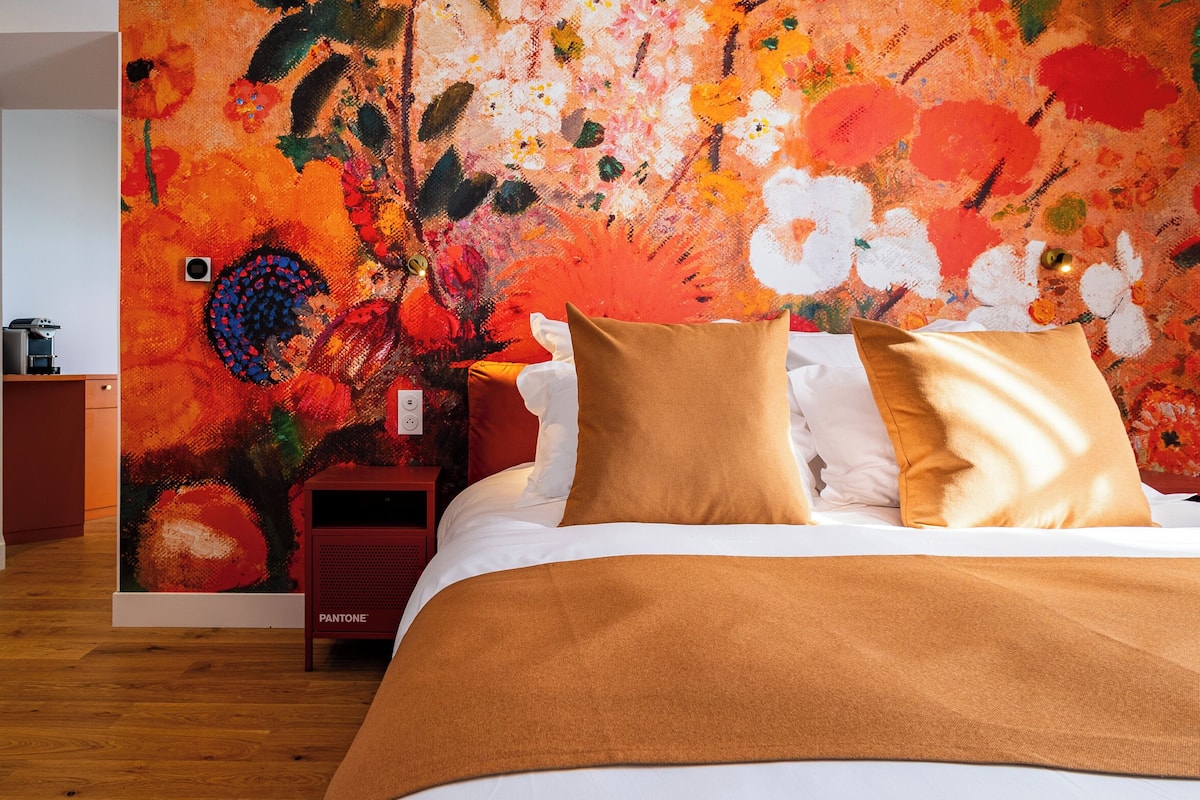 Ein Schlafzimmer mit einer großen Blumenwand, einem Bett mit weißer und brauner Bettwäsche sowie einem roten Nachttisch mit Lampe.