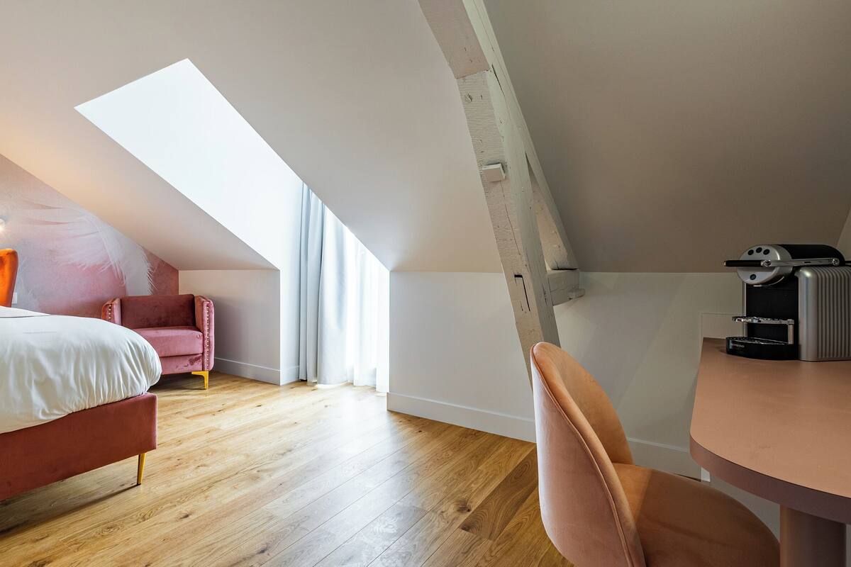Ein modernes Schlafzimmer mit schrägem Dach, einem Bett, einem rosa Sessel und einer Kaffeemaschine.
