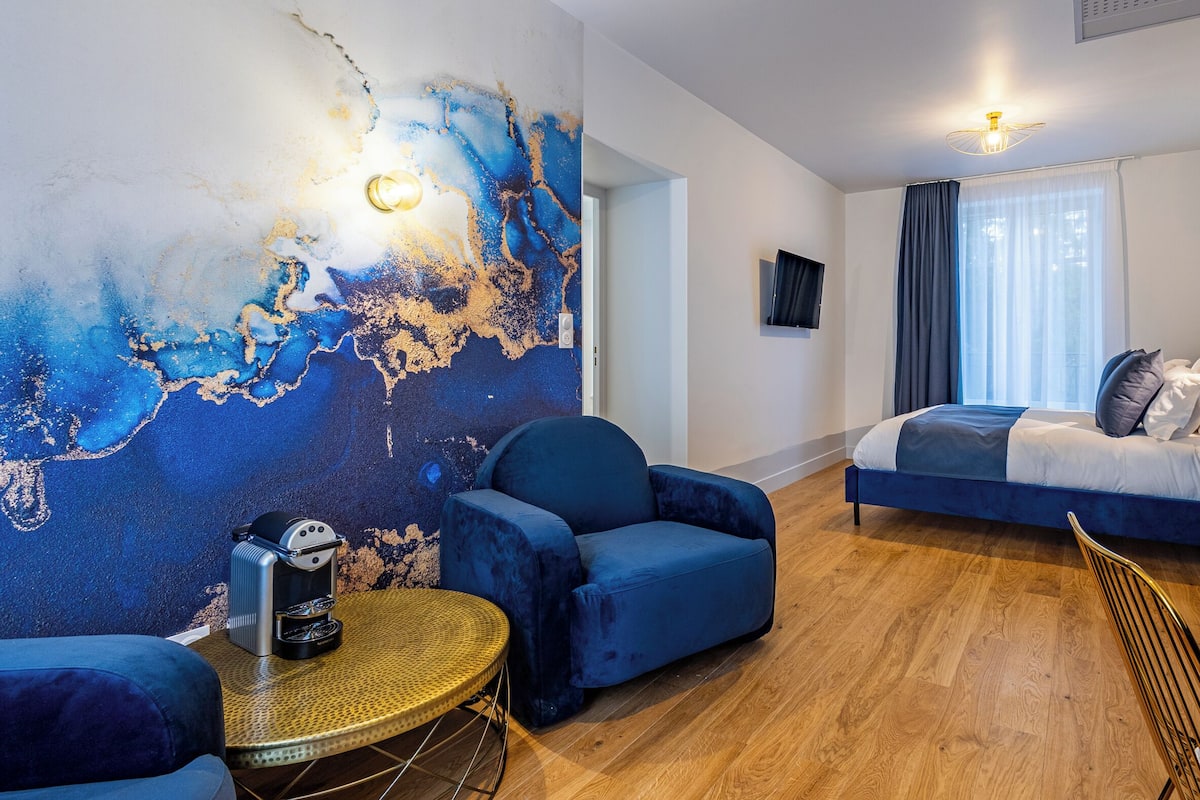 Ein modernes Schlafzimmer mit einem großen Bett, einer Sitzecke mit einem Sessel und einem kleinen runden Tisch sowie einer Wand mit einem blauen, abstrakten Wandbild.