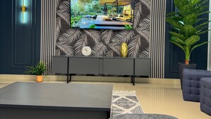 A modern living room with a flat-screen TV mounted on a wall with a leaf-patterned design. There is a sofa, a coffee table, and a potted plant.