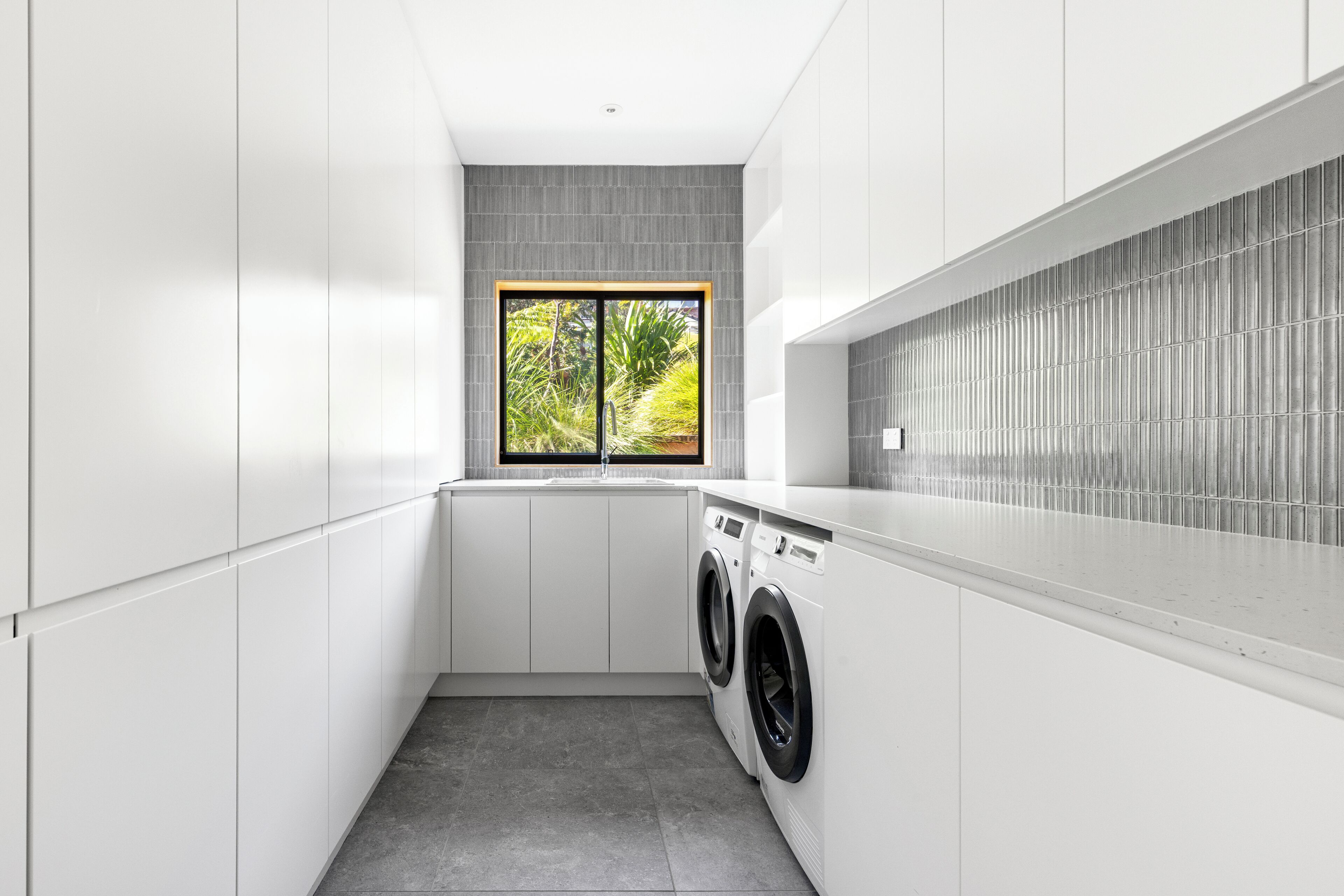 A modern laundry room with a built-in washer and dryer, white cabinetry, and a window with a view of greenery.