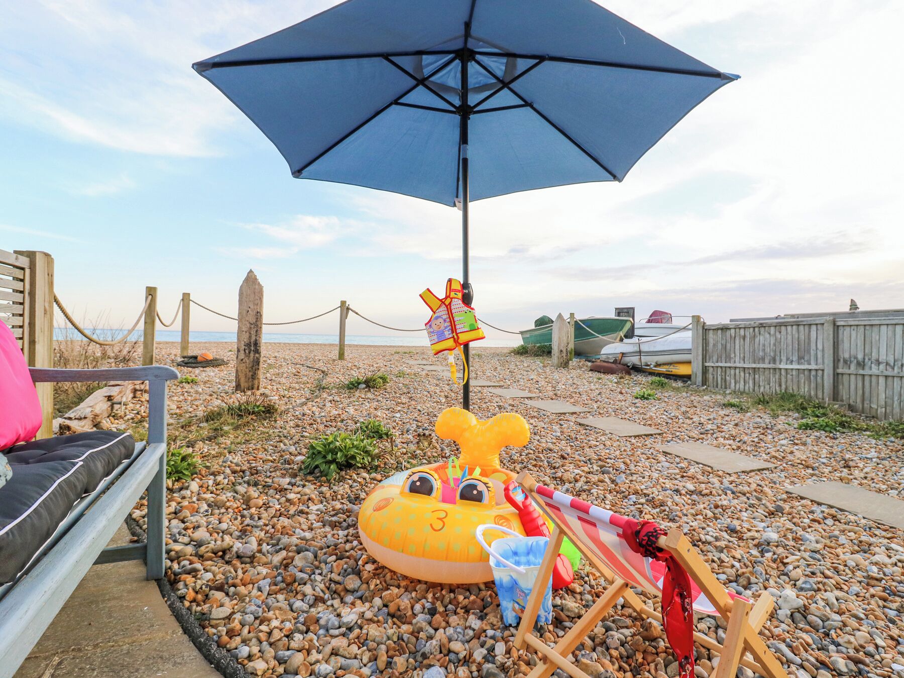 A beach area with a wooden fence, a beach chair, a large blue umbrella, and a yellow inflatable toy.