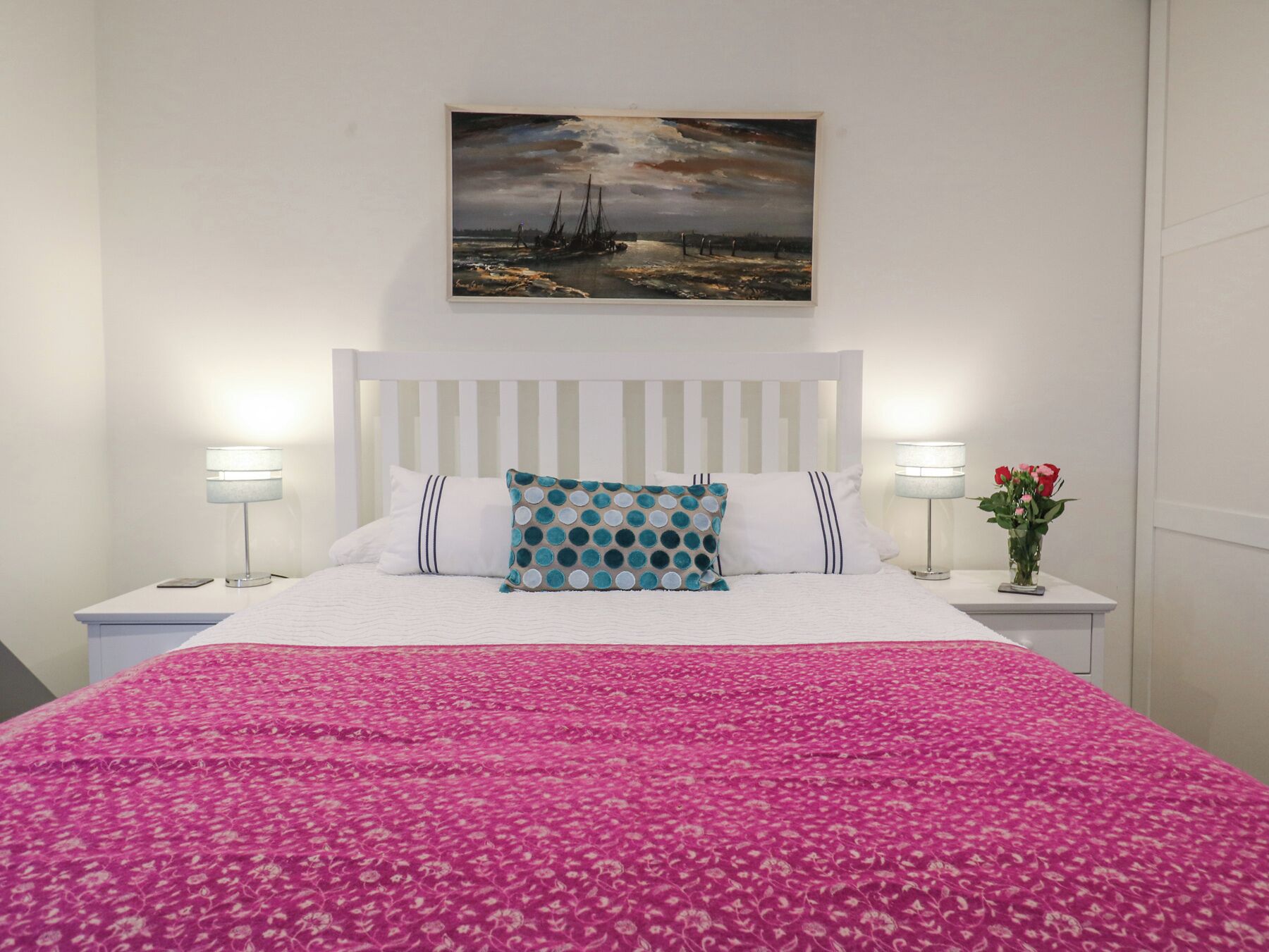 A bed with a pink floral bedspread, white pillows, and a teal pillow with circular patterns. There is a painting of a seascape above the bed.