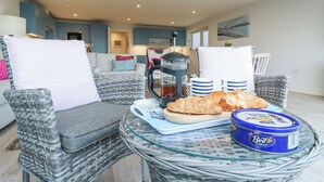 Un coin salon extérieur accueillant, avec des fauteuils en osier, une table où sont disposés des croissants et une cafetière, ainsi qu’une boîte bleue contenant du beurre.