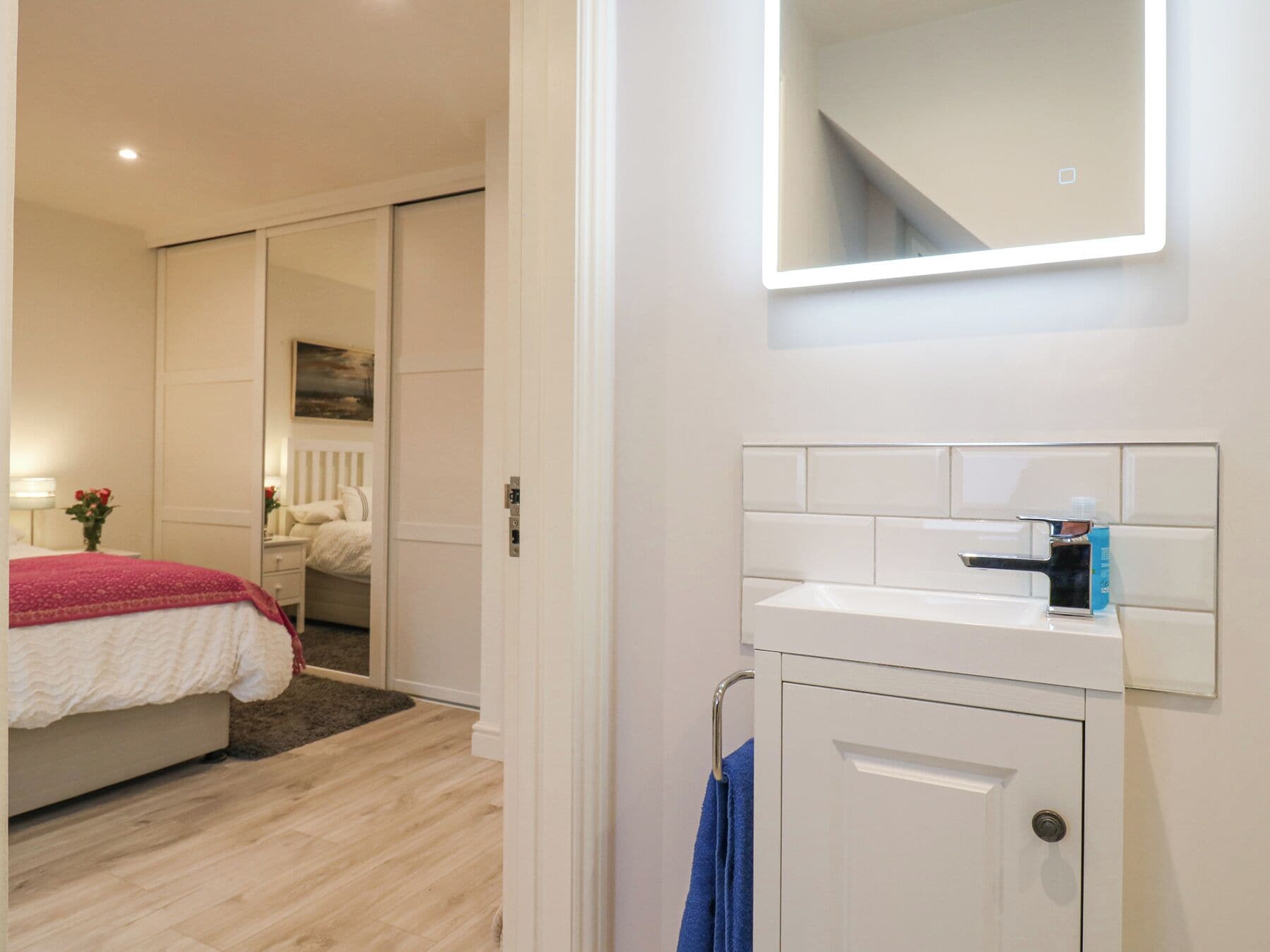 A bedroom with a bed, a nightstand, a dresser, and a bathroom with a mirror, sink, and towel rack.