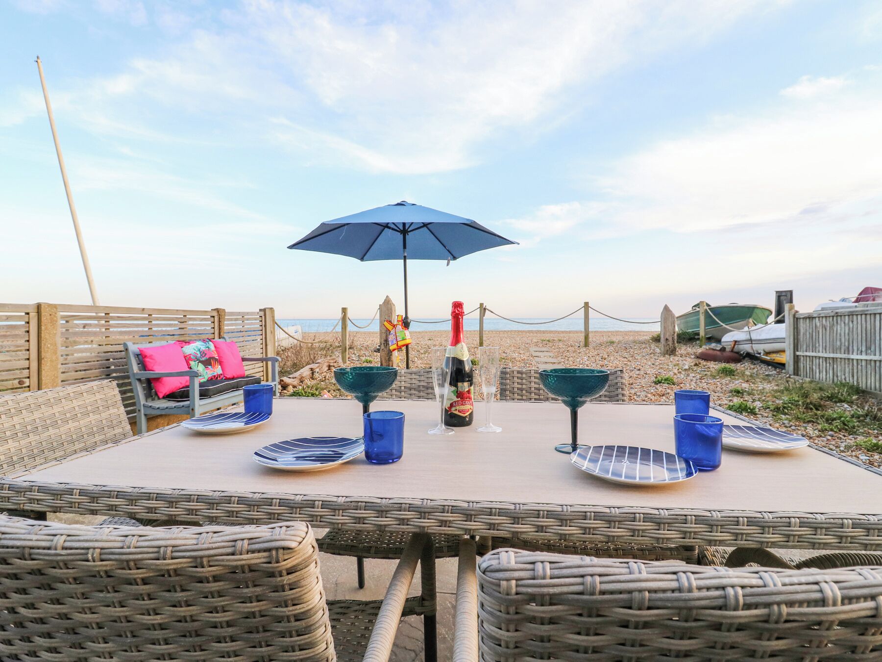 A beachside table set with wicker chairs, a bottle of champagne, and a blue umbrella.