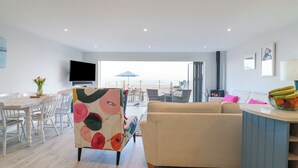 Un salon moderne avec un canapé beige, un fauteuil coloré, une table à manger avec des chaises, un téléviseur à écran plat et une vue sur la plage grâce à de grandes fenêtres.