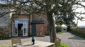 A wooden table with a bottle and glasses, set outdoors with a tree and a building in the background. - The Roost (Carlisle)