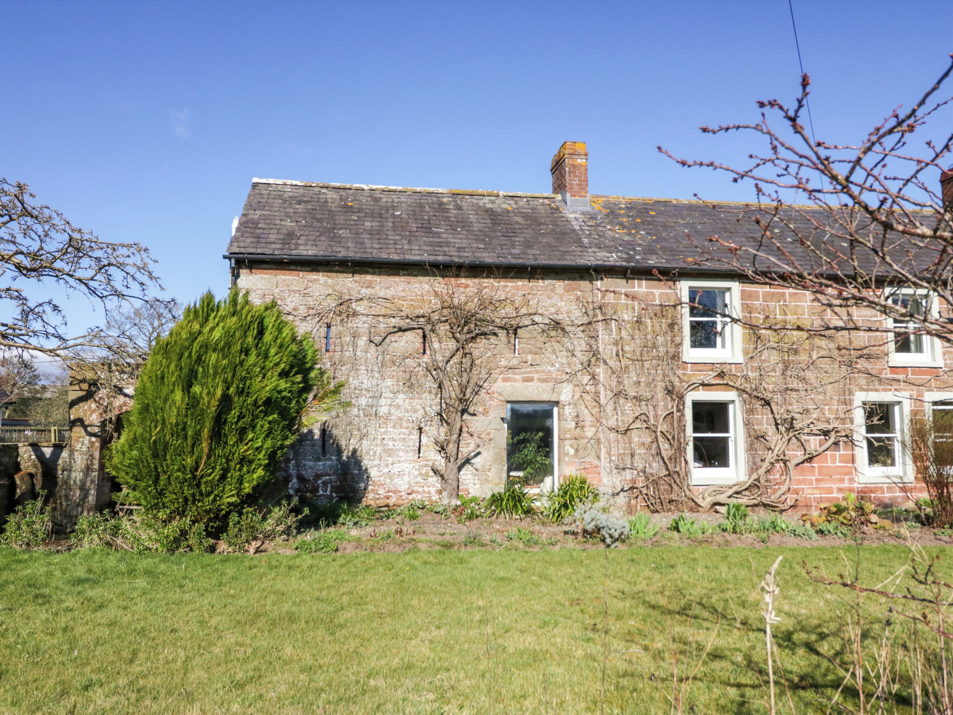 A stone house with a sloped roof, a chimney, and a small garden area with grass and a tree.