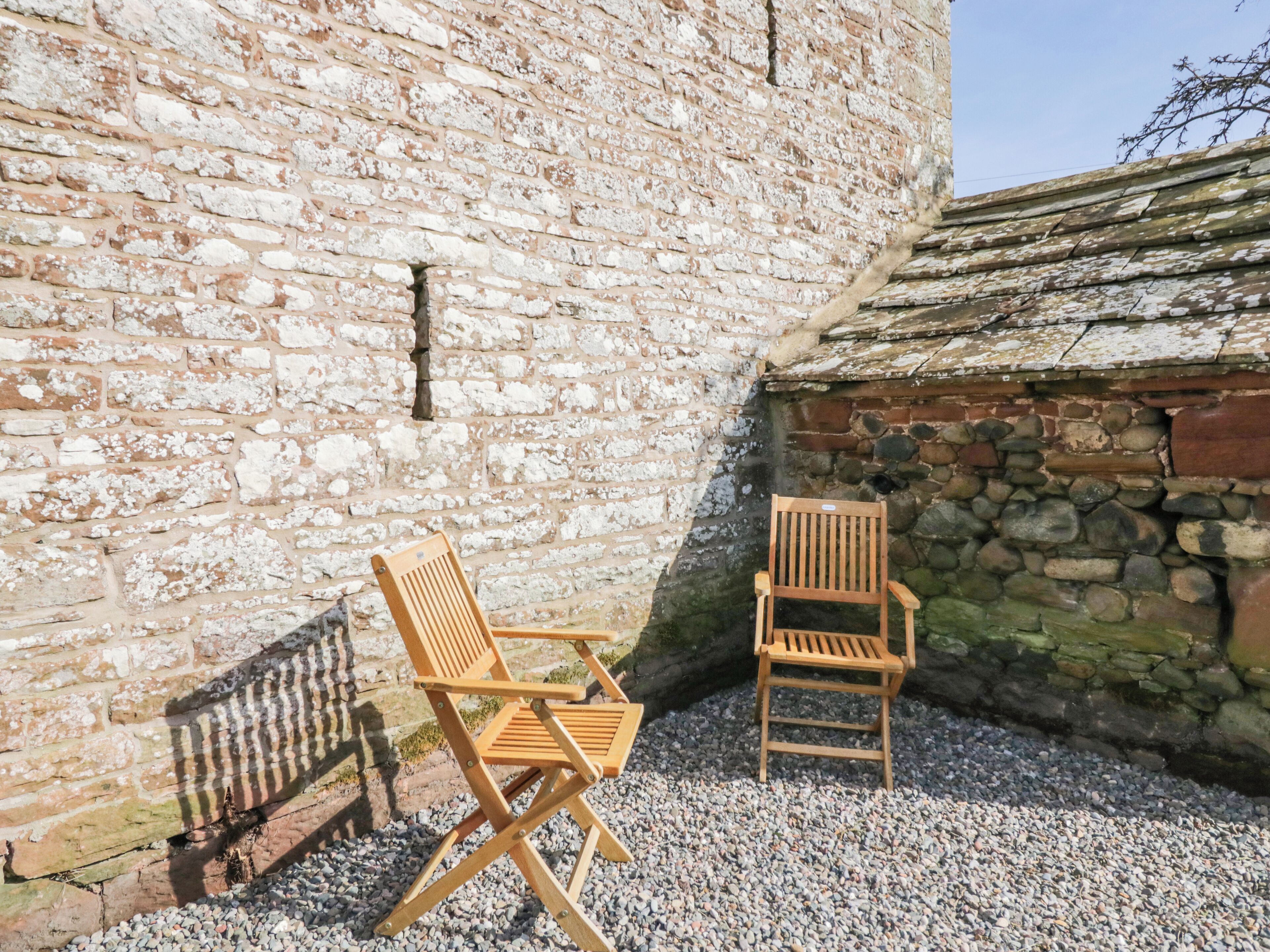 Two wooden chairs positioned on gravel in front of a stone wall with a tiled roof.