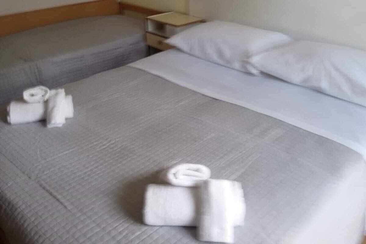 Habitación de hotel con dos camas, cada una con ropa de cama y almohadas blancas. Hay toallas enrolladas con cuidado sobre las camas. Se ve una mesita de noche con una lámpara y una pequeña mesa.