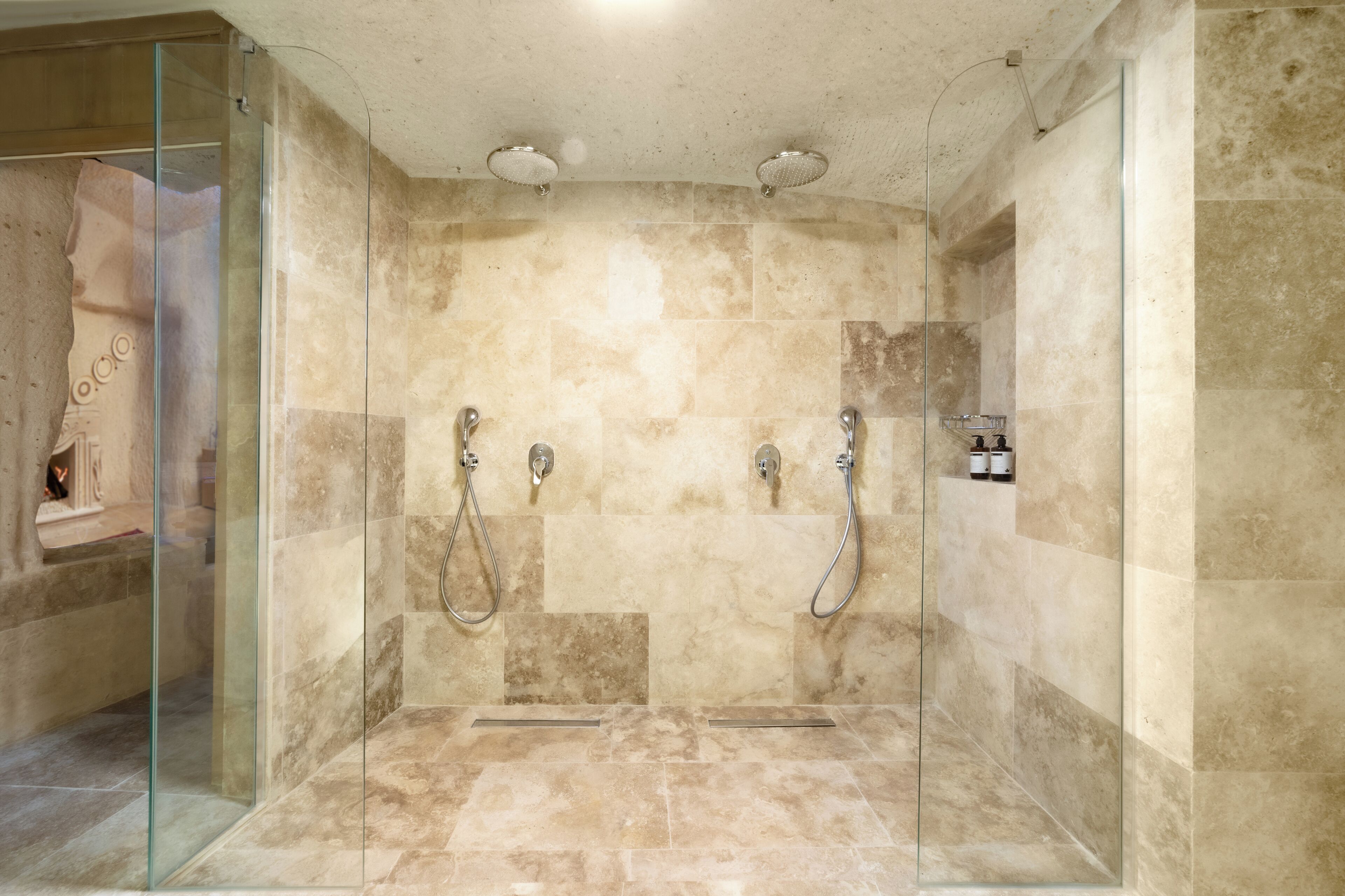 A modern shower with glass doors, beige tiles, and two handheld showerheads.