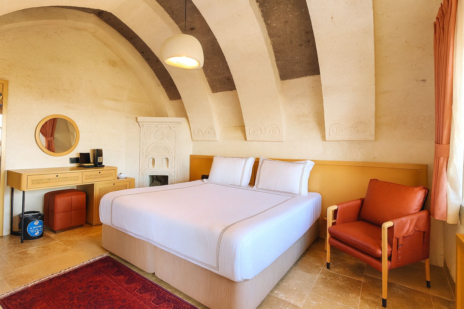 A bedroom with a large bed, a desk with a mirror, a red leather chair, and a fireplace.