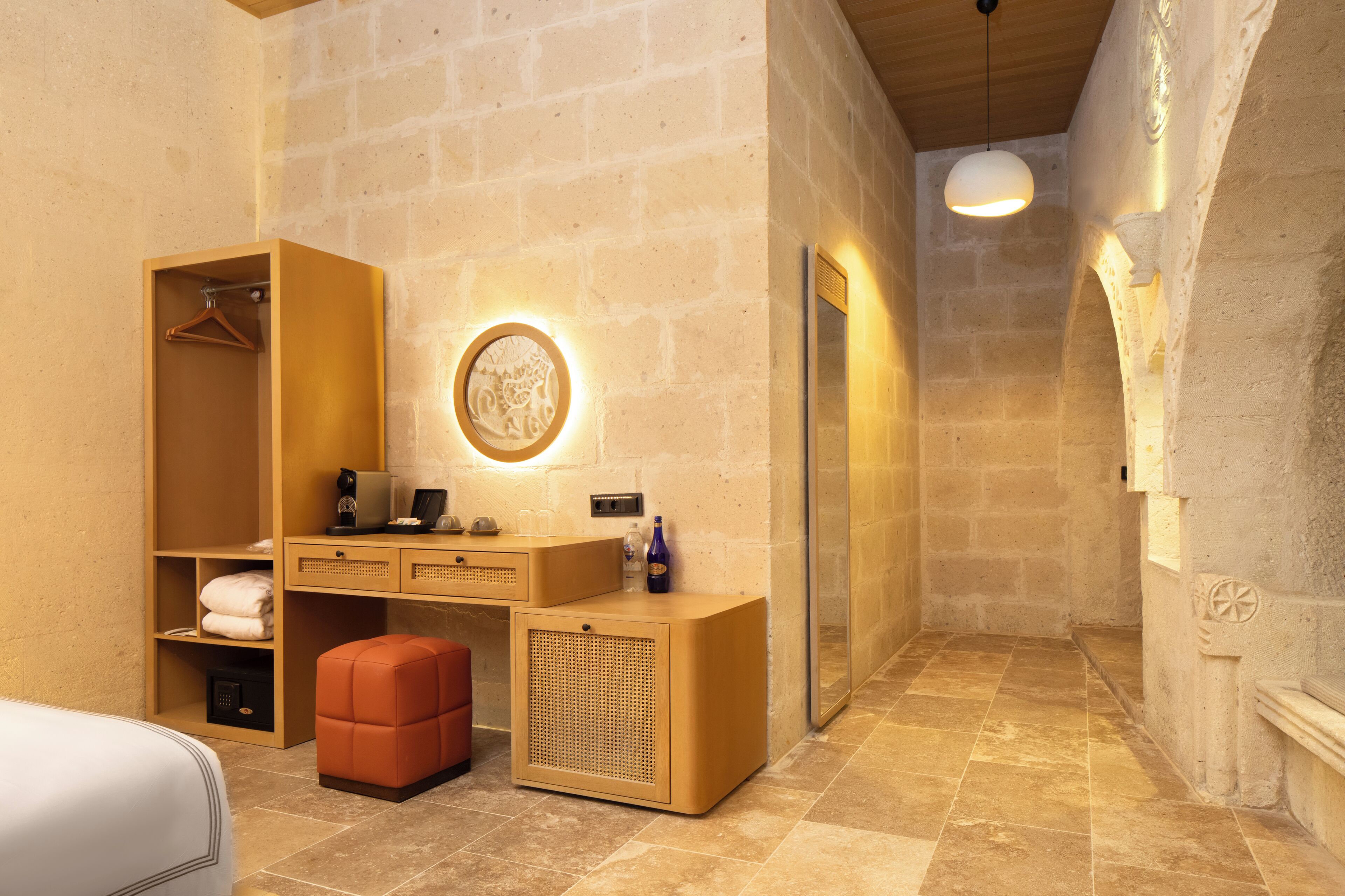 A hotel room with a wooden wardrobe, a desk with a mirror, a round wall light, and a bench.