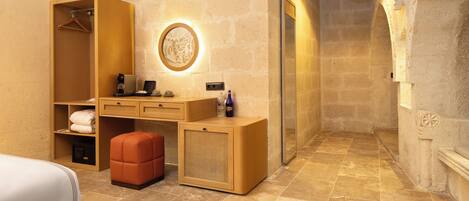 A hotel room with a wooden wardrobe, a desk with a mirror, a round wall light, and a bench.