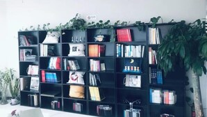 A black bookshelf filled with books and decorative items, surrounded by green plants. - Shanyehong Hotel (Beijing)