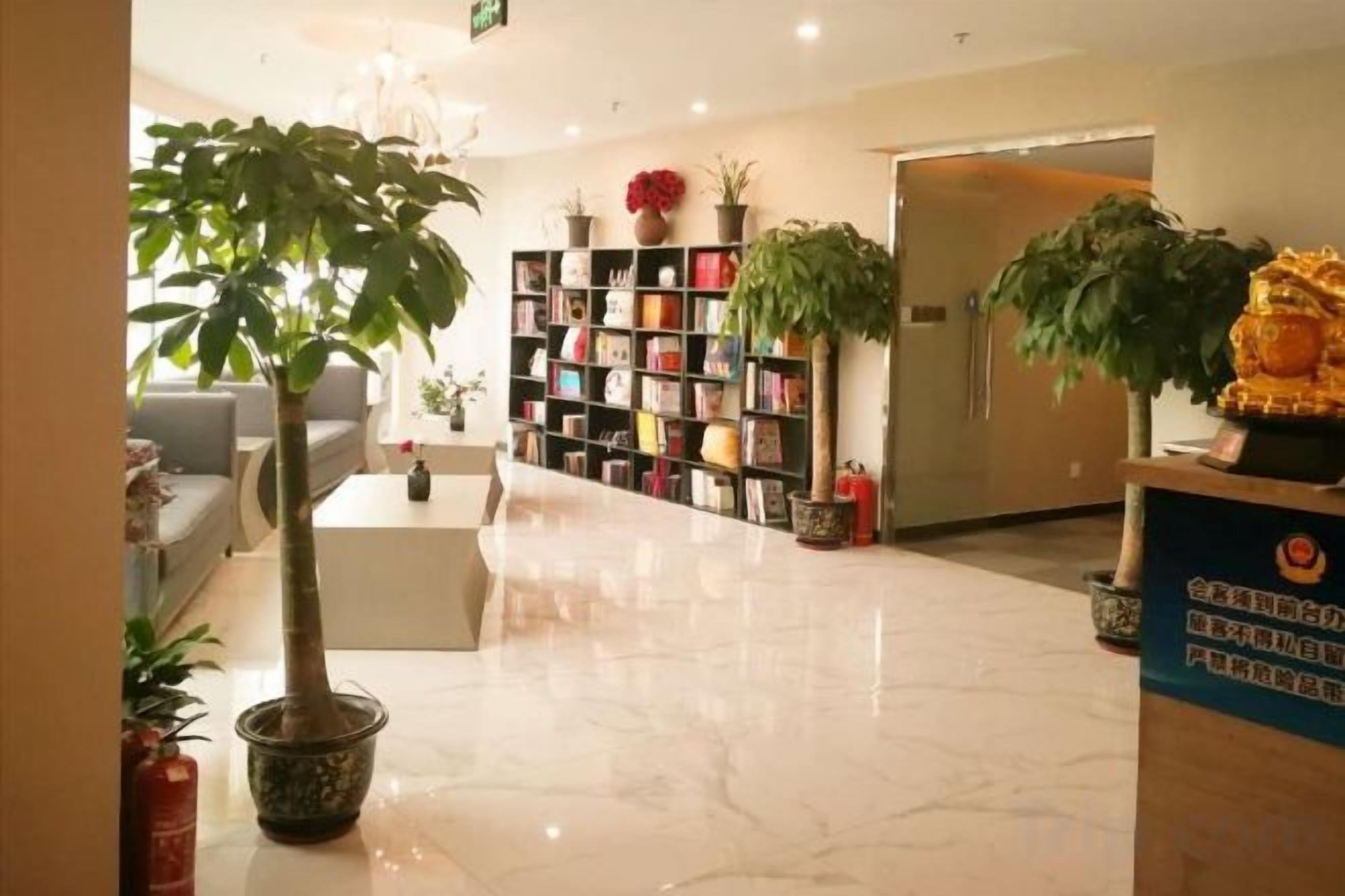 Un vestíbulo de hotel con una recepción, una zona de estar, una estantería y plantas en macetas.