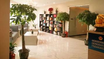Un vestíbulo de hotel con una recepción, una zona de estar, una estantería y plantas en macetas.