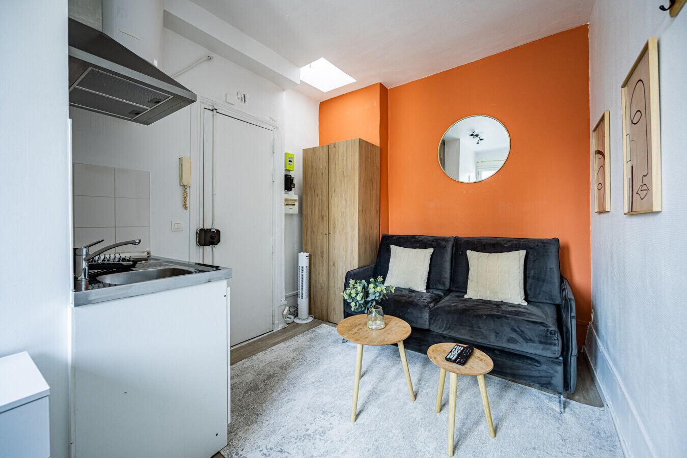 A compact living space with a kitchenette, a black sofa, and a round mirror on the orange wall.