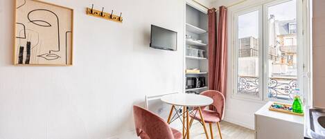 Una habitación moderna con una mesa redonda, dos sillas rosas, un televisor, una estantería y una ventana con vistas a edificios.