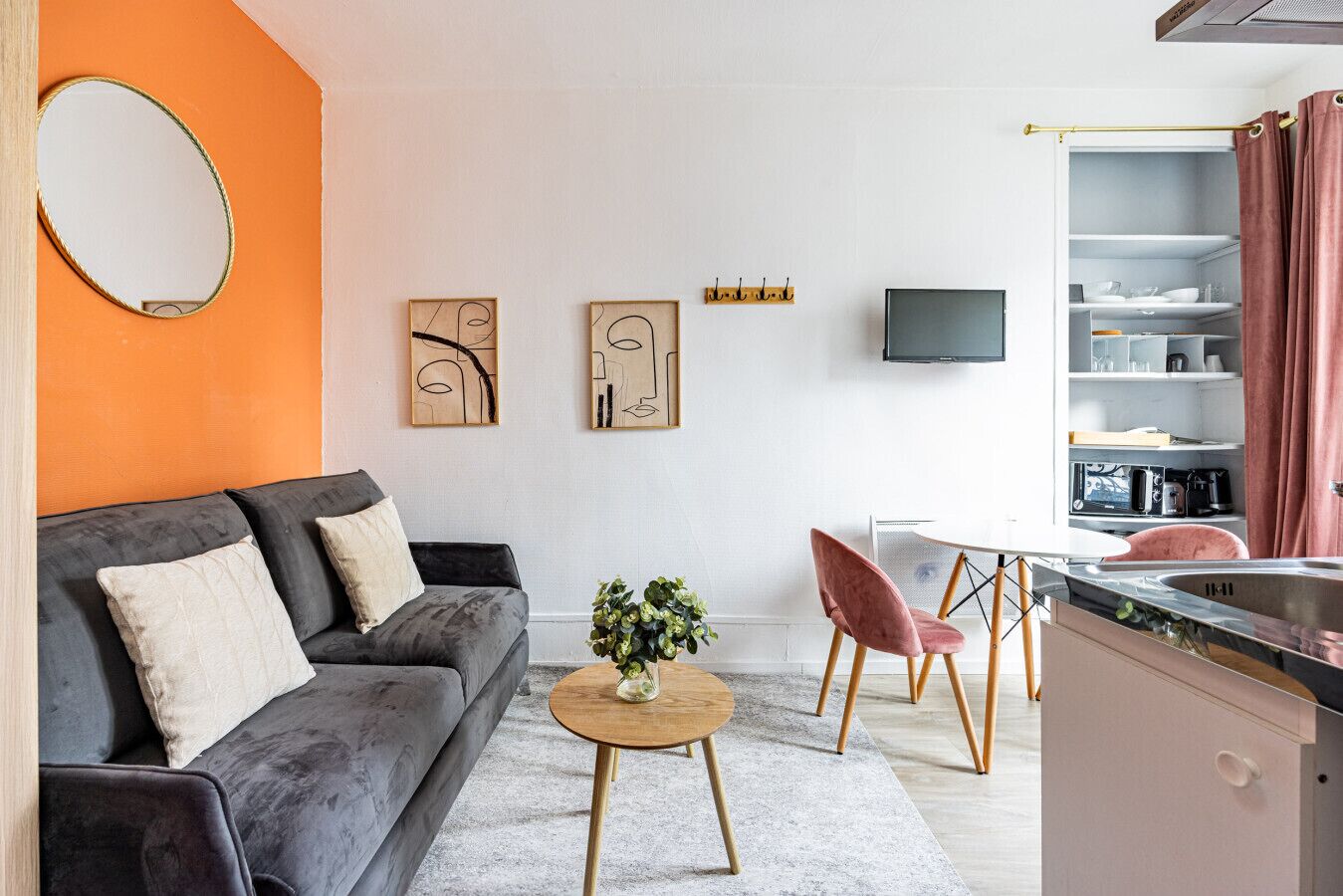 A modern living room with a grey sofa, a small round table with a plant, and a kitchen area with a sink and cabinets.