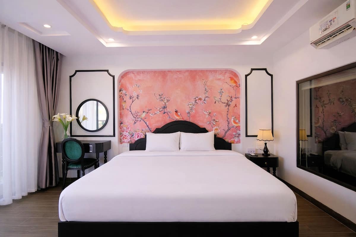 A hotel room with a large bed, a desk with a chair, a mirror, and a wall with a floral painting.