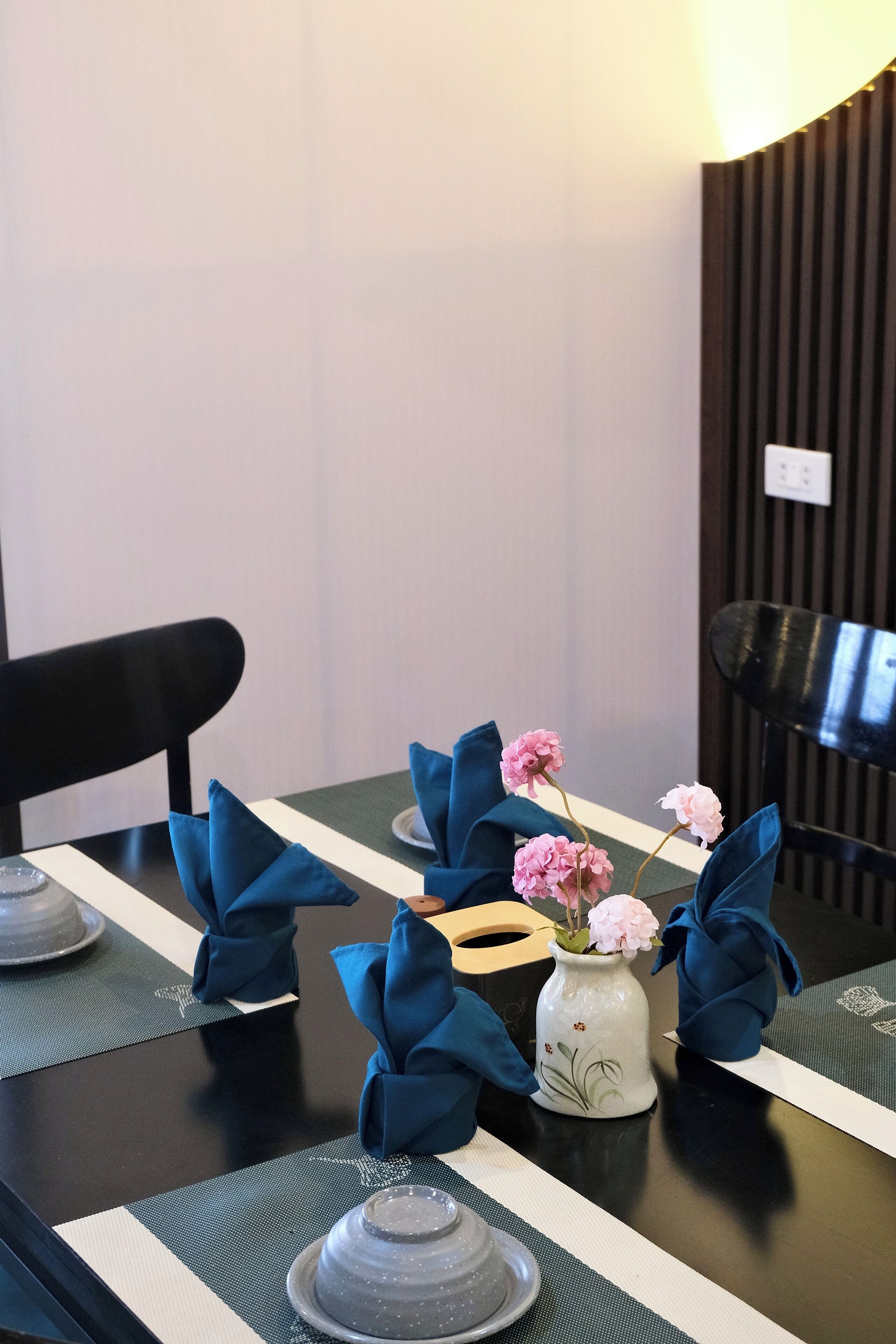 A dining table set with blue napkins, a vase with pink flowers, and a plate with a glass.