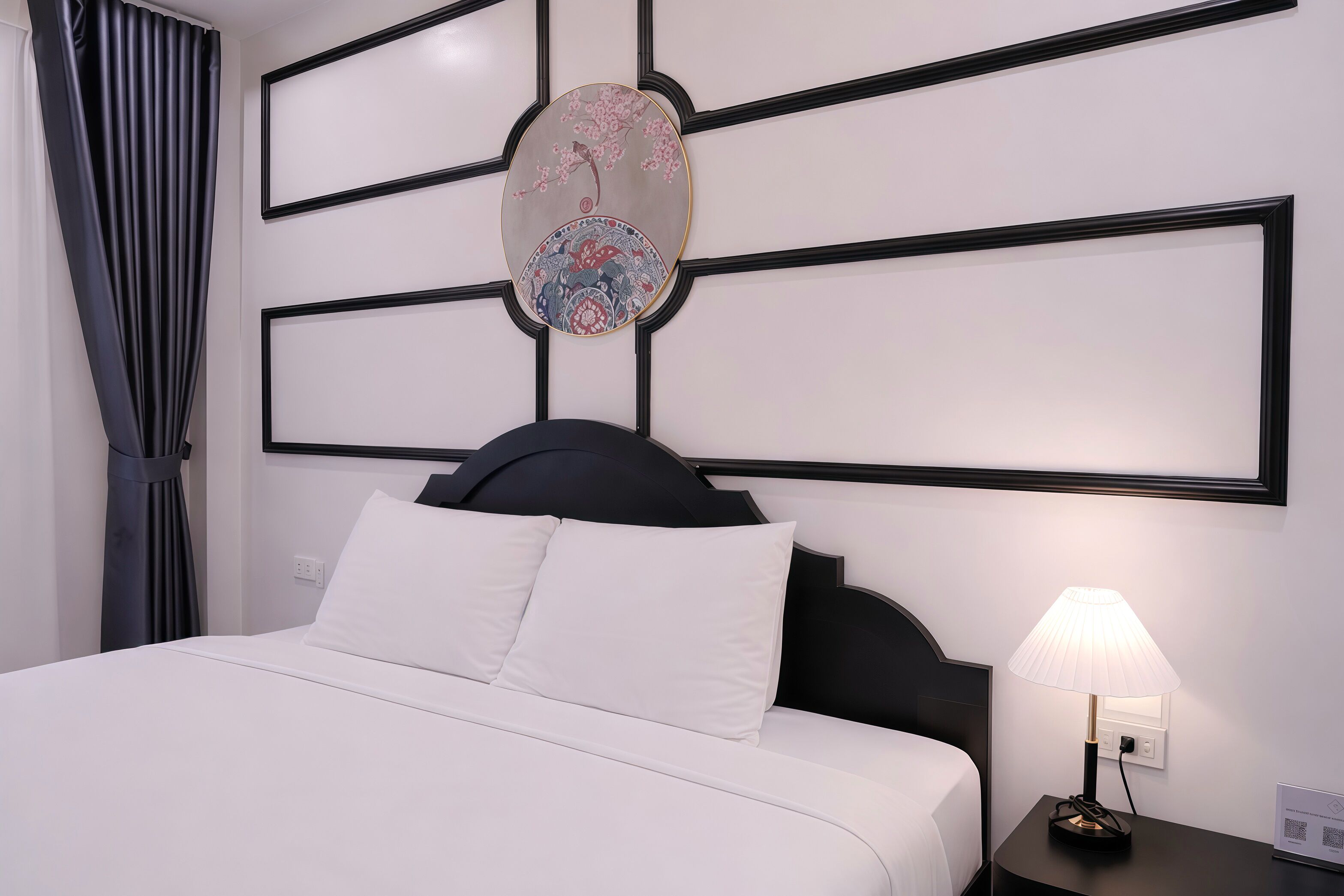 Quarto de hotel com cama, mesa de cabeceira com abajur, uma obra de arte na parede e janela com cortinas.
