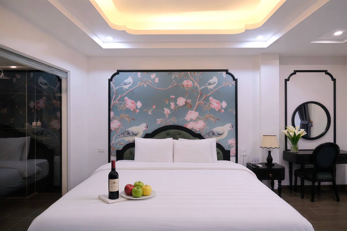 A hotel room with a large bed, a desk with a chair, a mirror, and a wall with floral wallpaper.