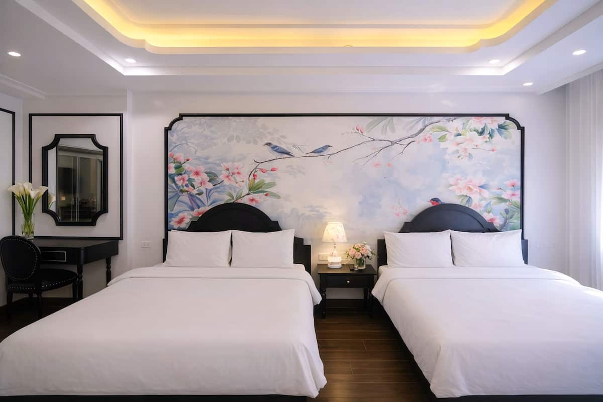 A hotel room with two beds, a desk, a chair, a nightstand with a lamp, and a wall painting.