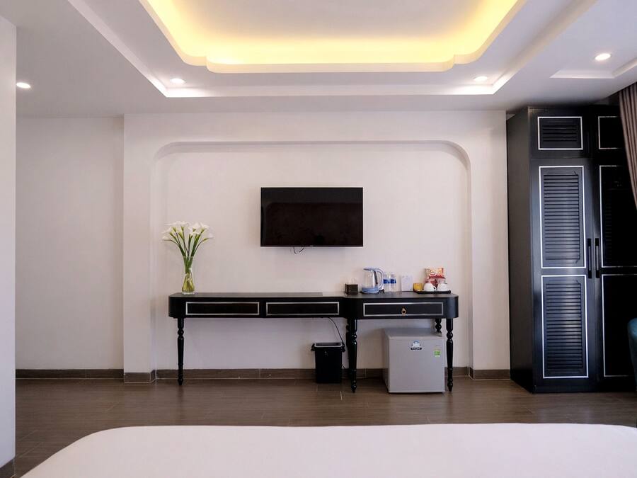 Una habitación de hotel moderna con una televisión de pantalla plana montada en la pared, un escritorio pequeño con una cafetera, un cubo de basura y un refrigerador miniatura.