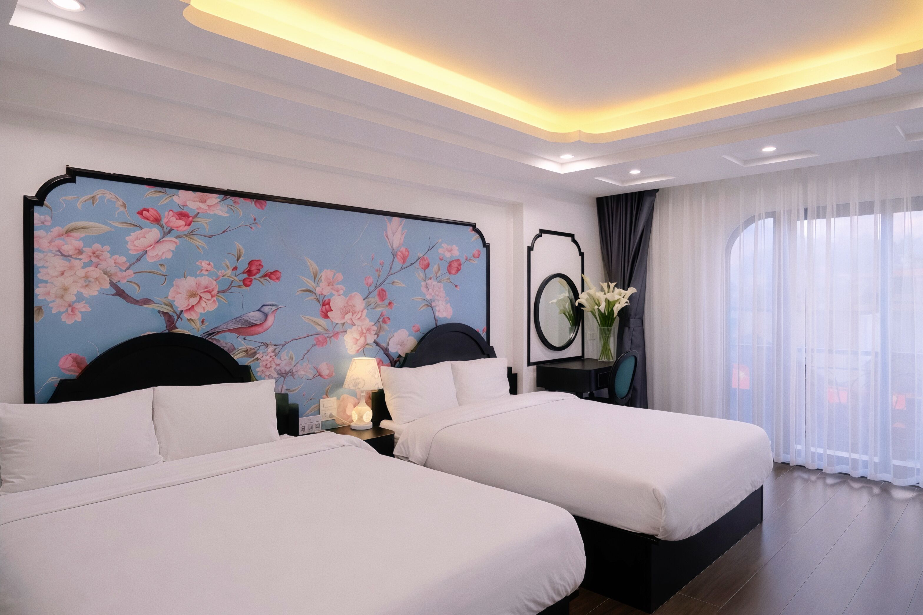 A hotel room with two beds, a floral mural on the wall, a nightstand with a lamp, and a view of the outdoors through sheer curtains.