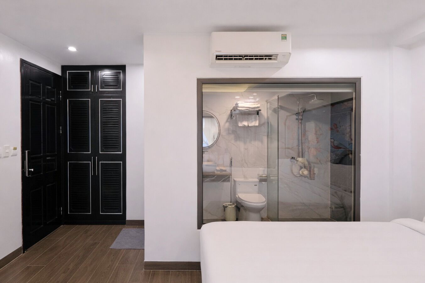 Habitación de hotel moderna con una cama grande, un vestidor con puertas negras, un baño con cabina de ducha de vidrio y un equipo de aire acondicionado sobre la ducha.