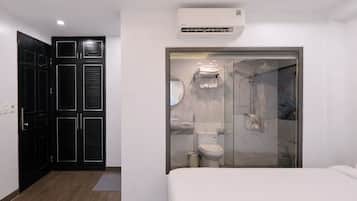 A modern hotel room with a large bed, a walk-in closet with black doors, a bathroom with a glass shower enclosure, and an air conditioning unit above the shower.