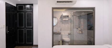 Habitación de hotel moderna con una cama grande, un vestidor con puertas negras, un baño con cabina de ducha de vidrio y un equipo de aire acondicionado sobre la ducha.